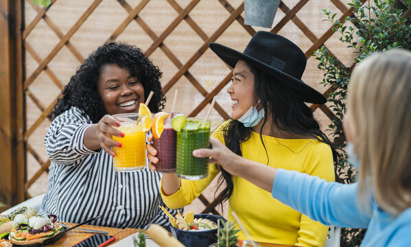 Multiracial friends toasting with fruits fresh smoothies in coffee brunch bar during corona virus outbreak