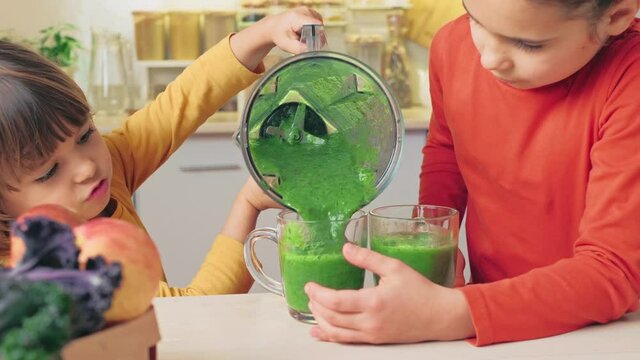 Happy Kids pour a green smoothie cocktail from a blender into glasses. Children learn to cook a fresh and healthy breakfast. Cheerful vegan family enjoying cooking activity, portrait. Healthy Habits 