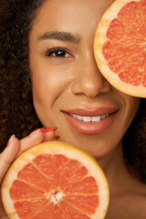 Face closeup of lovely mixed race young woman smiling at camera, posing with grapefruit cut in half