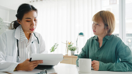 Young Asia female doctor in white medical uniform using clipboard is delivering great news talk discuss results or symptoms with girl patient sitting at desk in health clinic or hospital office.