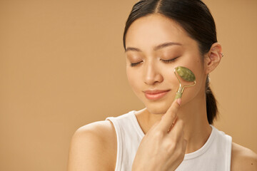 Facial beauty treatment. Beautiful young woman looking relaxed while getting massage face using jade roller isolated over beige background