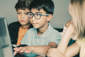 Asian boy typing on laptop keyboard and classmates sitting at table, watching him and doing task together. Pupils using computers during study. Childhood, communication and digital education concept