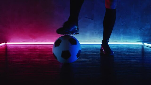 Soccer player rolls a ball on floor by his leg. Football workout training concept. Athlete sportsman with the ball in dark studio with magenta blue lights. Sport. Shot on Blackmagic ProRes. Bet