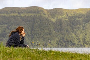 Naklejka na ściany i meble Girl sitting in grass, by Sete Cidades lake, enjoying the landscape, peacefully.