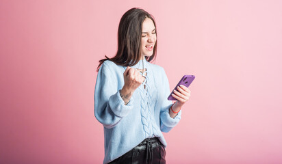 Portrait of a brunette on a pink background who uses a smartphone and shows the gesture of the winner