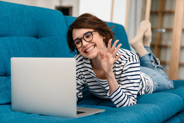 Happy nice woman using laptop and waving hand while lying on couch