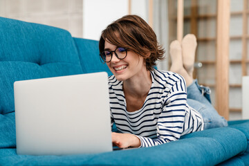 Happy beautiful woman in eyeglasses using laptop while lying on couch