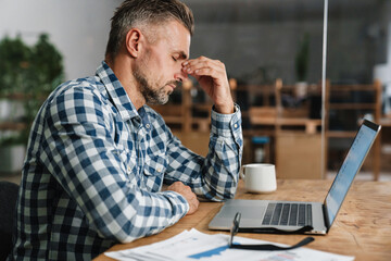 Unhappy grey man with headache working with laptop in office