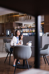 happy freelancer looking away while sitting near laptop in restaurant, blurred foreground