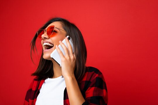 Closeup photo of attractive positive laughing young brunet woman wearing stylish red shirt white t-shirt and red sunglasses isolated over red background communicating on mobile phone looking up