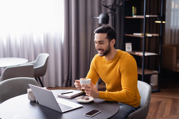 excited arabian freelancer laughing while holding cup of coffee near laptop in restaurant