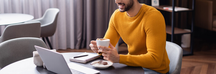 cropped view of arabian freelancer holding cup of coffee near laptop, banner