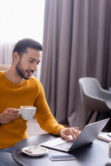 smiling arabian freelancer holding cup of coffee while typing on laptop in restaurant