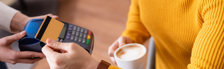 partial view of man holding cup of coffee and credit card near payment terminal in hands of waitress, blurred background, banner