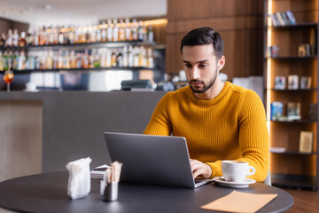 focused arabian freelancer working on laptop near cup of coffee in restaurant, blurred foreground