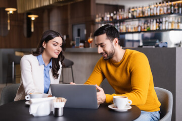 cheerful arabian freelancer pointing at laptop near smiling woman in restaurant, blurred foreground