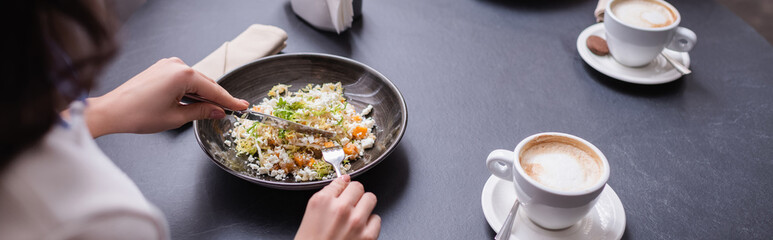 Cropped view of woman holding cutlery near salad and cup of cappuccino in restaurant, banner