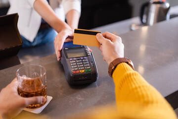 Cropped view of man paying with credit card near bartender and glass of whiskey on blurred foreground