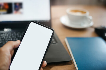 Selective focus on blank screen mobile in human hand with blur computer laptop and coffee cup on background