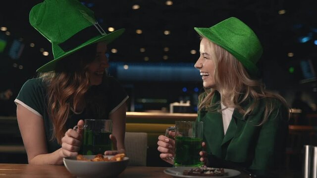 Women having fun drinking beer and laughing. Female friends in Irish hats celebrating Saint Patrick's Day in a pub. 
