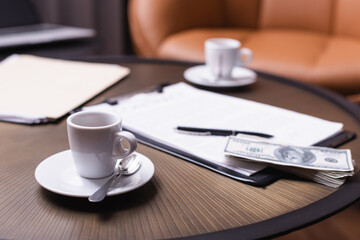 Cup of coffee near dollars and clipboard on blurred background on table