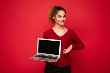 side profile portrait photo of beautiful charming fascinating young curly dark blond lady wearing red sweater standing isolated over red wall background holding computer laptop with empty copy space