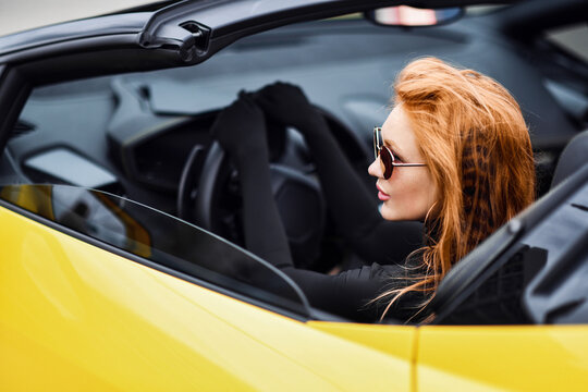 Portrait of rich sporty red-haired woman in black clothes, long gloves and sunglasses is sitting behind the wheel driving yellow premium sport car cabriolet convertible and looking aside