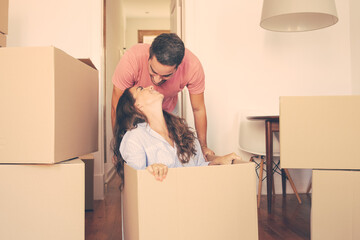 Obraz na płótnie Canvas Cheerful young man dragging box with his girlfriend inside and kissing her. Joyful family couple having fun while moving into new flat. New home concept