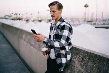 Handsome caucasian hipster guy in trend wear browsing web page on smartphone reading news on free time outdoors,20s young hipster guy blogging and sending mail via mobile phone holding coffee cup