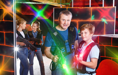 Portrait of glad positive teen boy and his father standing indoors with laser guns ready for lasertag game.