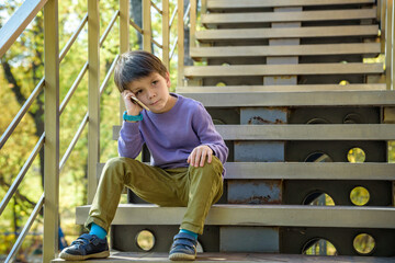 Closeup portrait, worried, sad young child, boy talking on phone to someone, looking unhappy, isolated outdoors background. Negative human emotions, facial expressions, feeling, reaction Communication