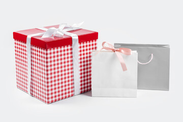 Red giftbox with white silk bow on top and paper shopping bags on light background. Gift boxes, surprise for holiday. Present, gift, shopping and sale concept