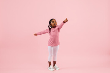 Pointing, showing. Childhood and dream about big and famous future. Pretty little girl isolated on coral pink studio background. Dreams, imagination, education, facial expression, emotions concept.