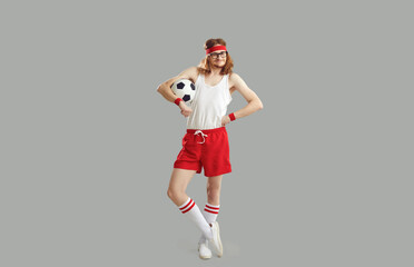 Full body happy satisfied nerdy man in glasses, headband, white tank top and red sports shorts standing akimbo, holding soccer ball and smiling isolated on grey background. Humorous football concept