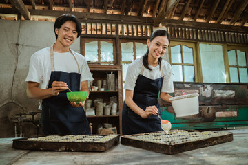 smiling man and woman wearing an apron looking at the camera while stirring the cake dough in a bowl near the cake tin on the table.