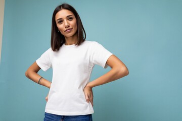 Photo of positive young beautiful brunette woman with sincere emotions wearing casual white t-shirt for mockup isolated over blue background with copy space - obrazy, fototapety, plakaty