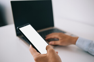 Crop man working on laptop and messaging on smartphone in office