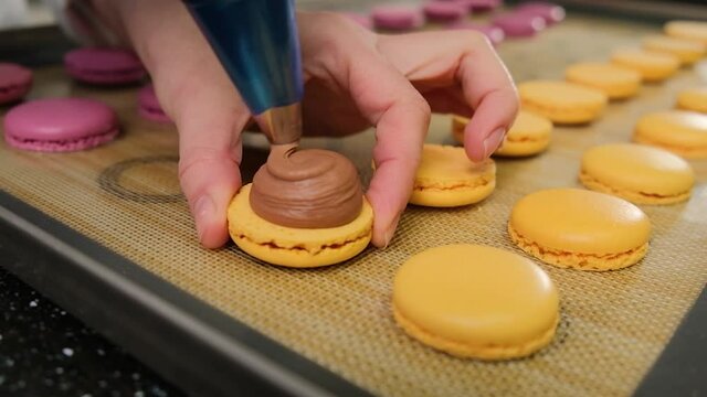 Pastry chef is filling cream chocolate macarons with pastry bag, close up.