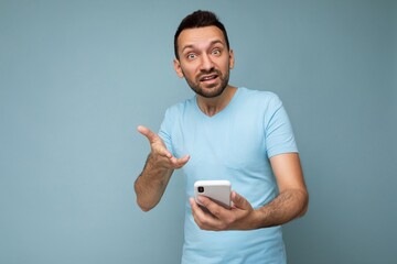 Photo of angry handsome young man with beard wearing everyday blue t-shirt isolated over blue background holding and using mobile phone communication online on the internet looking at camera and