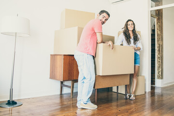 Fototapeta na wymiar Young couple moving into new flat, leaning on cardboard boxes and furniture indoors, looking at camera and smiling. Full length. New home or property buying concept