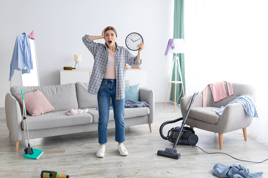 Shocked young lady with clock standing in messy room after party, too late to clean apartment before parents return