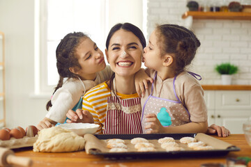Baking and cooking with children at home concept. Two small daughters kissing young smiling happy mother during baking sweet bicsuits together in kitchen at home