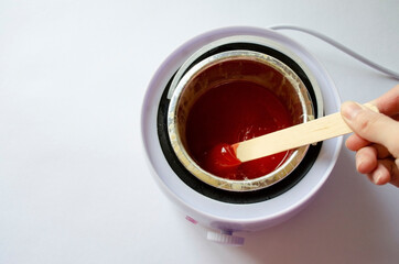 red depilatory wax melted in a wax melt. hand holding a stick with wax