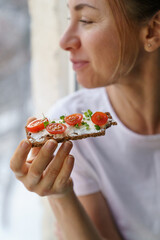 Happy woman eating rye crisp bread with creamy vegetarian cheese tofu, cherry tomato and rucola micro greens, sitting at home and looking at window. Healthy food, gluten free, diet concept. 