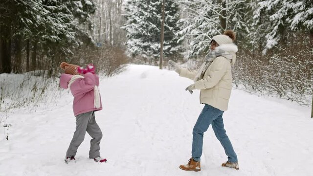 Long side view of happy Asian mom and daughter wearing puffer jackets, hats with pompoms and scarves smiling, having fun, playing snowballs in white winter forest