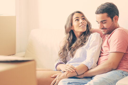 Happy Relaxed Young Couple Enjoying New Home, Relaxing On Couch Among Carton Boxes, Looking Around. Moving Or Property Buying Concept