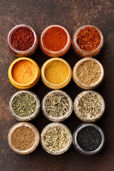 Set of various multicolored spices in jars over dark brown concrete background. Top view, close up, flat lay. Food background, ingredients for cooking, assorted condiment