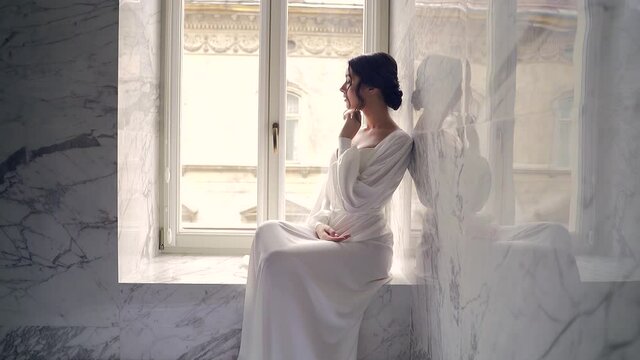 elegant bride in the morning in a dressing gown. young attractive woman in white lingerie in a luxury bathroom interior sitting on the windowsill near the window. looking seductively into the camera.