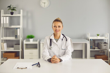 Portrait of friendly smiling doctor in white lab coat. Happy young woman who works as physician, general practitioner or healthcare consultant sitting at desk in her office at clinic or hospital