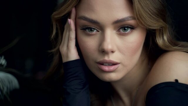 Close-up portrait of a young luxurious sexy woman looking at the camera on a dark background indoors. Face of professional fashion model with make-up miss with blue eyes. Film grain